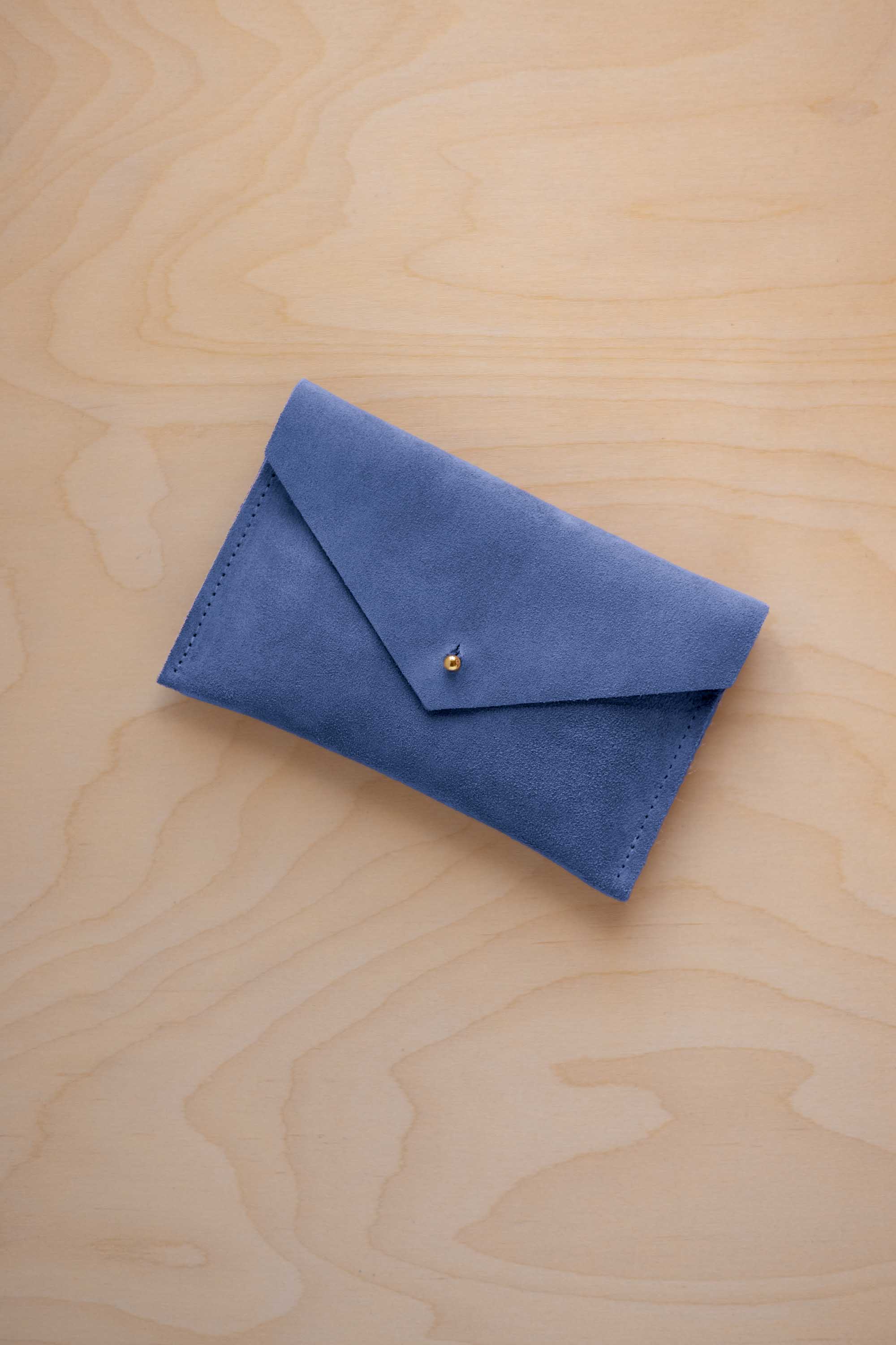 A small cobalt blue coloured suede envelope-style pouch with a gold button, resting on a light wood grain surface.
