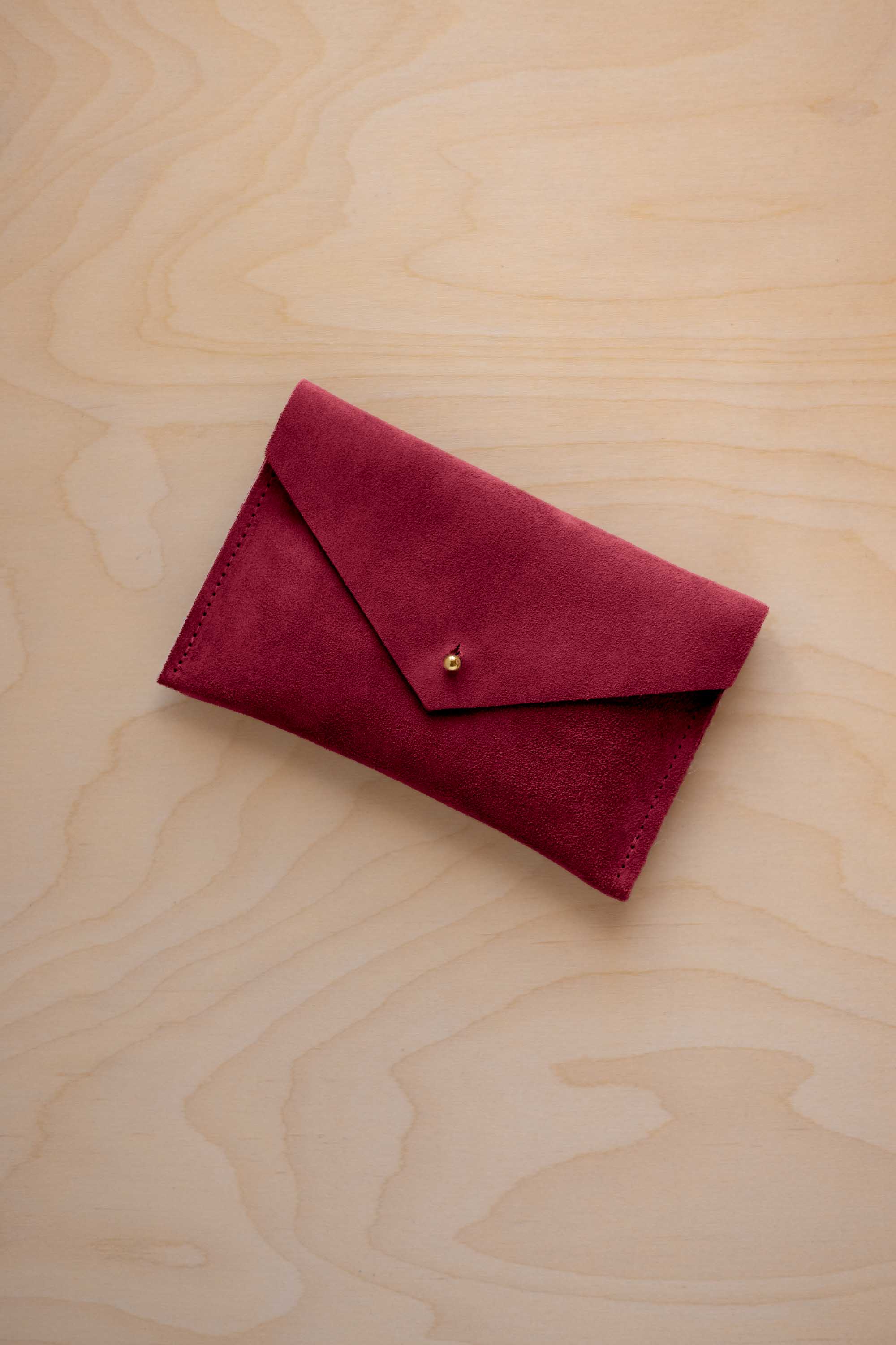 A small crimson red coloured suede envelope-style pouch with a gold button, resting on a light wood grain surface.