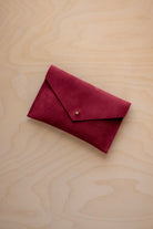 A small crimson red coloured suede envelope-style pouch with a gold button, resting on a light wood grain surface.