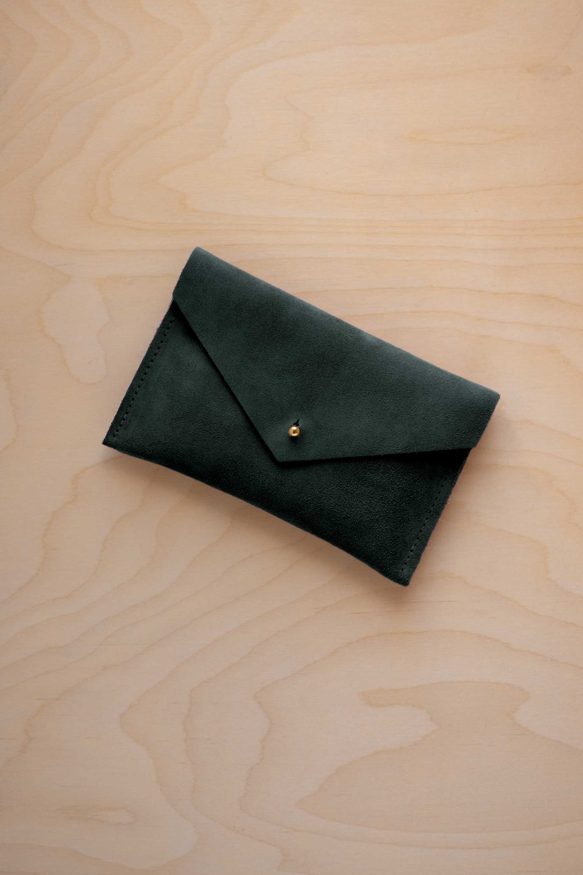 A small deep forest green coloured suede envelope-style pouch with a gold button, resting on a light wood grain surface.