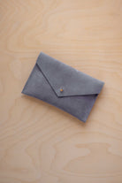 A small dusky blue coloured suede envelope-style pouch with a gold button, resting on a light wood grain surface.