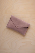 A small dusty mauve coloured suede envelope-style pouch with a gold button, resting on a light wood grain surface.