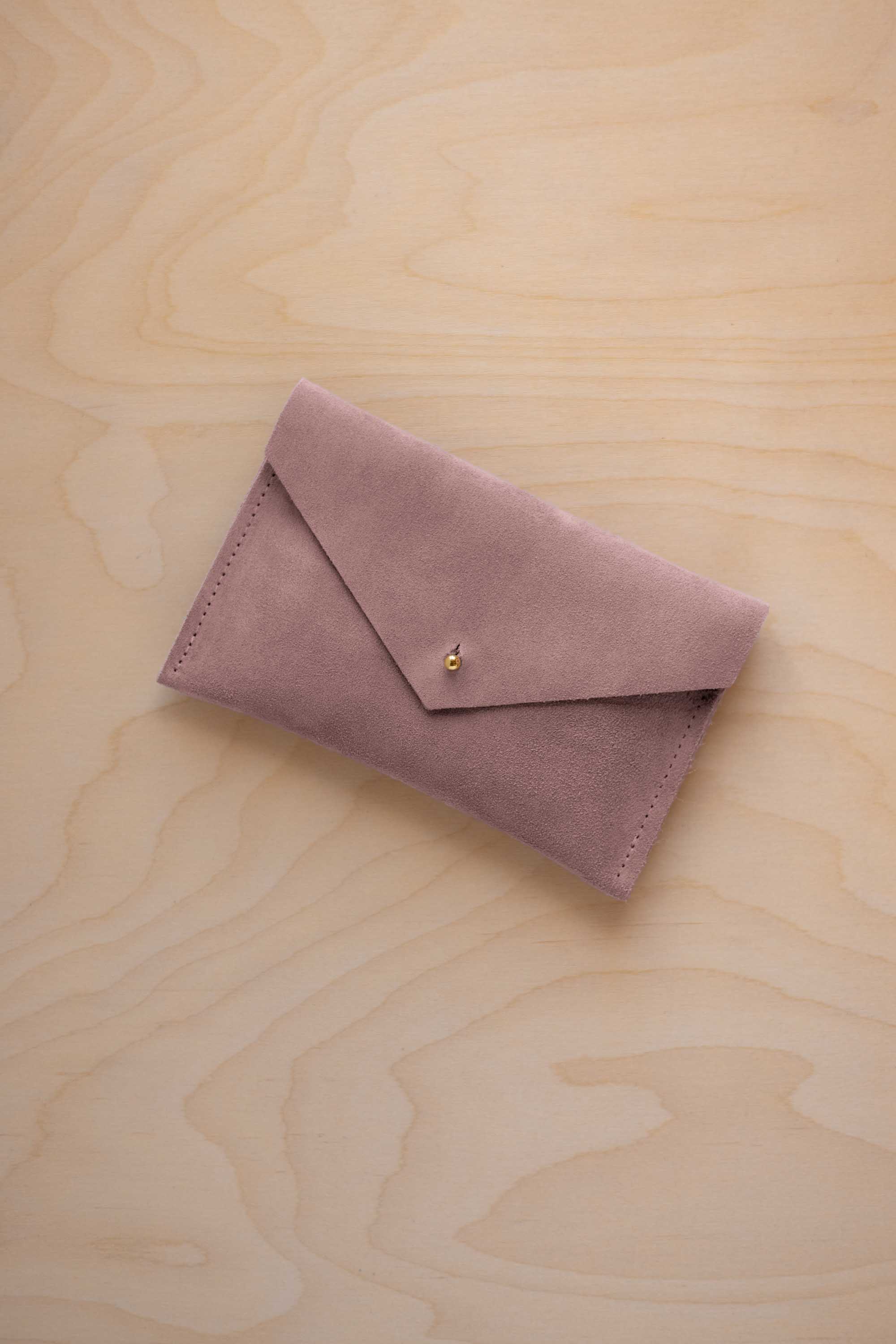 A small dusty mauve coloured suede envelope-style pouch with a gold button, resting on a light wood grain surface.