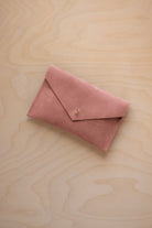 A small dusty rose coloured suede envelope-style pouch with a gold button, resting on a light wood grain surface.