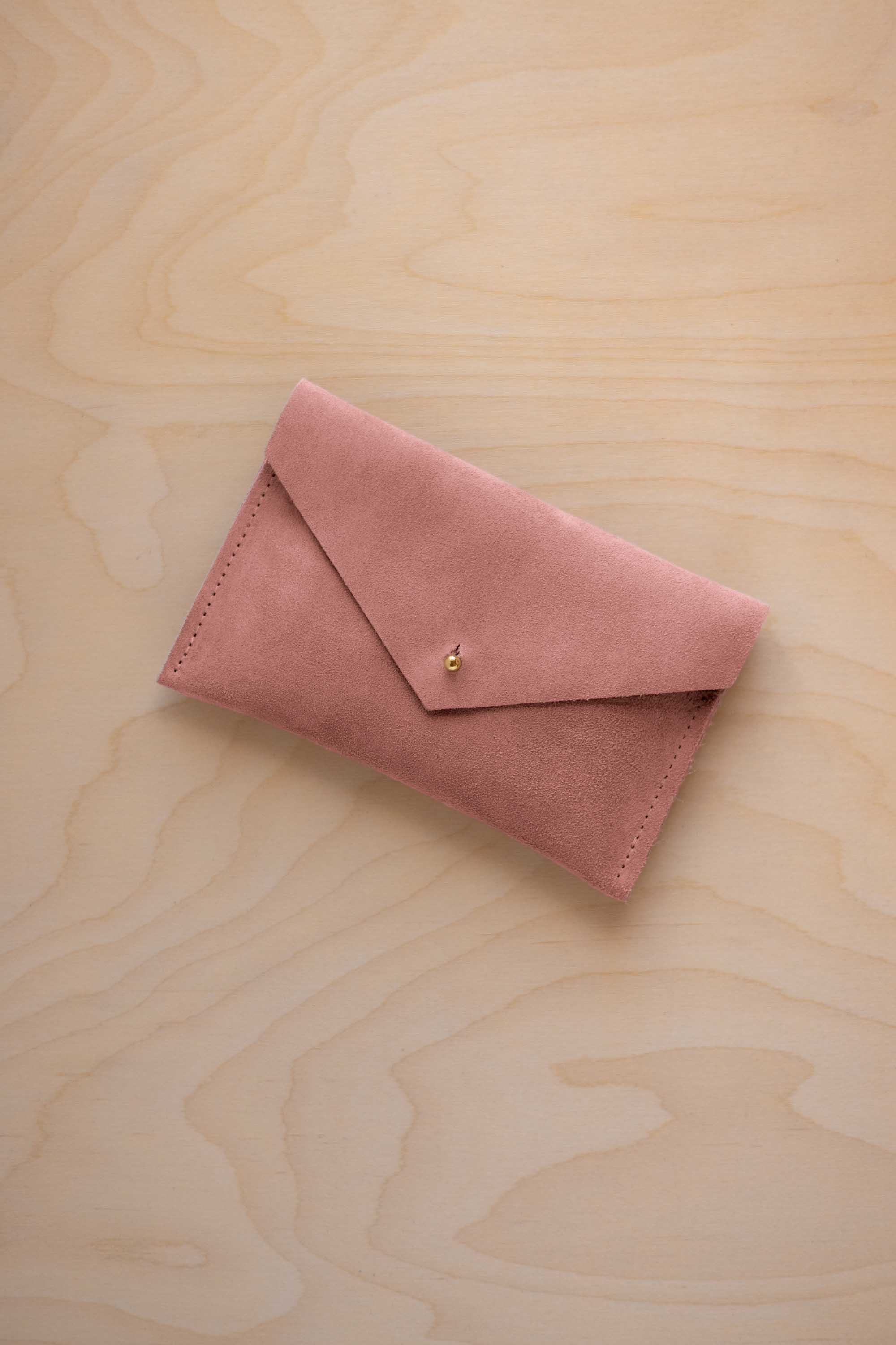 A small dusty rose coloured suede envelope-style pouch with a gold button, resting on a light wood grain surface.