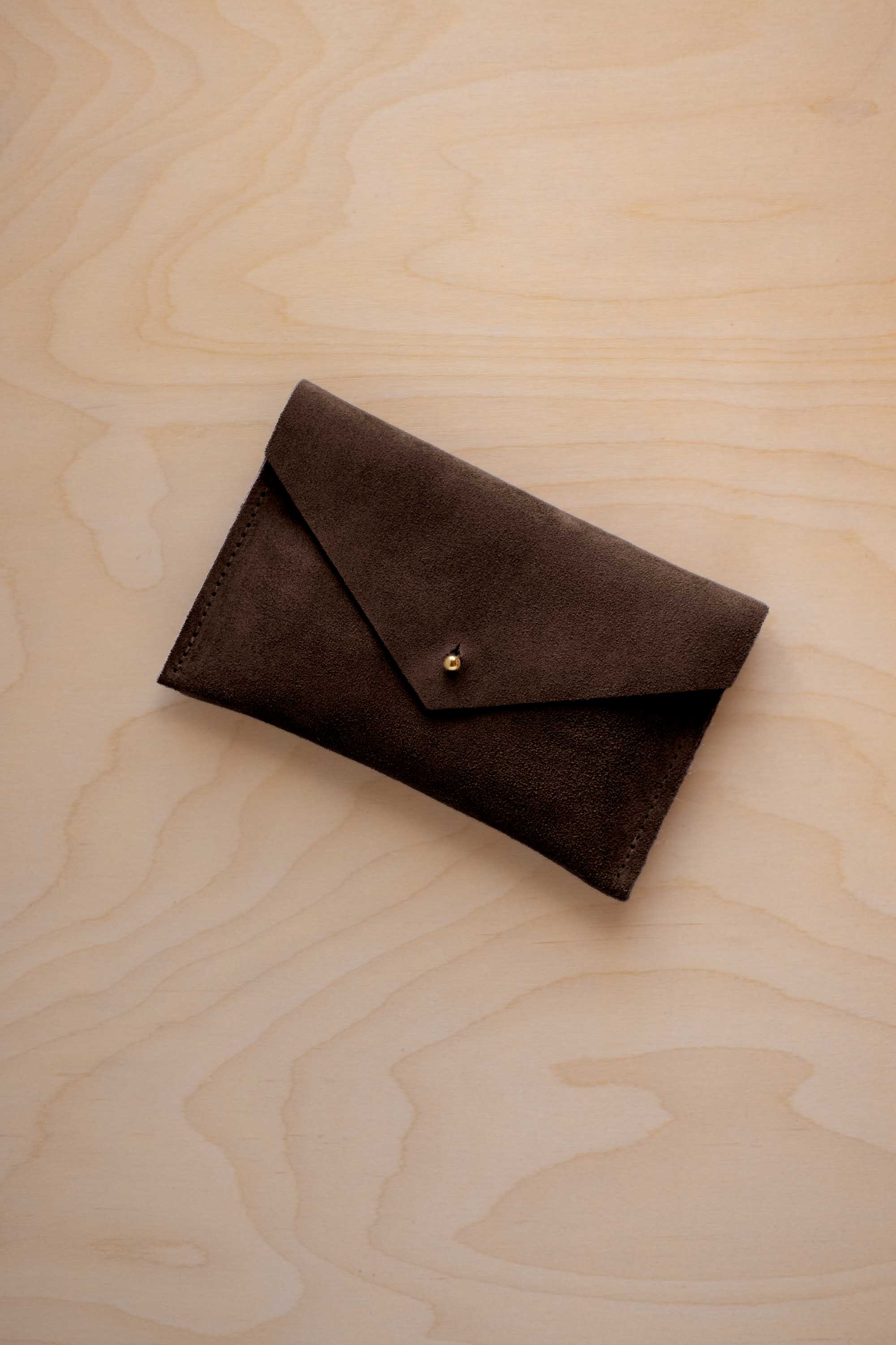 A small espresso brown coloured suede envelope-style pouch with a gold button, resting on a light wood grain surface.