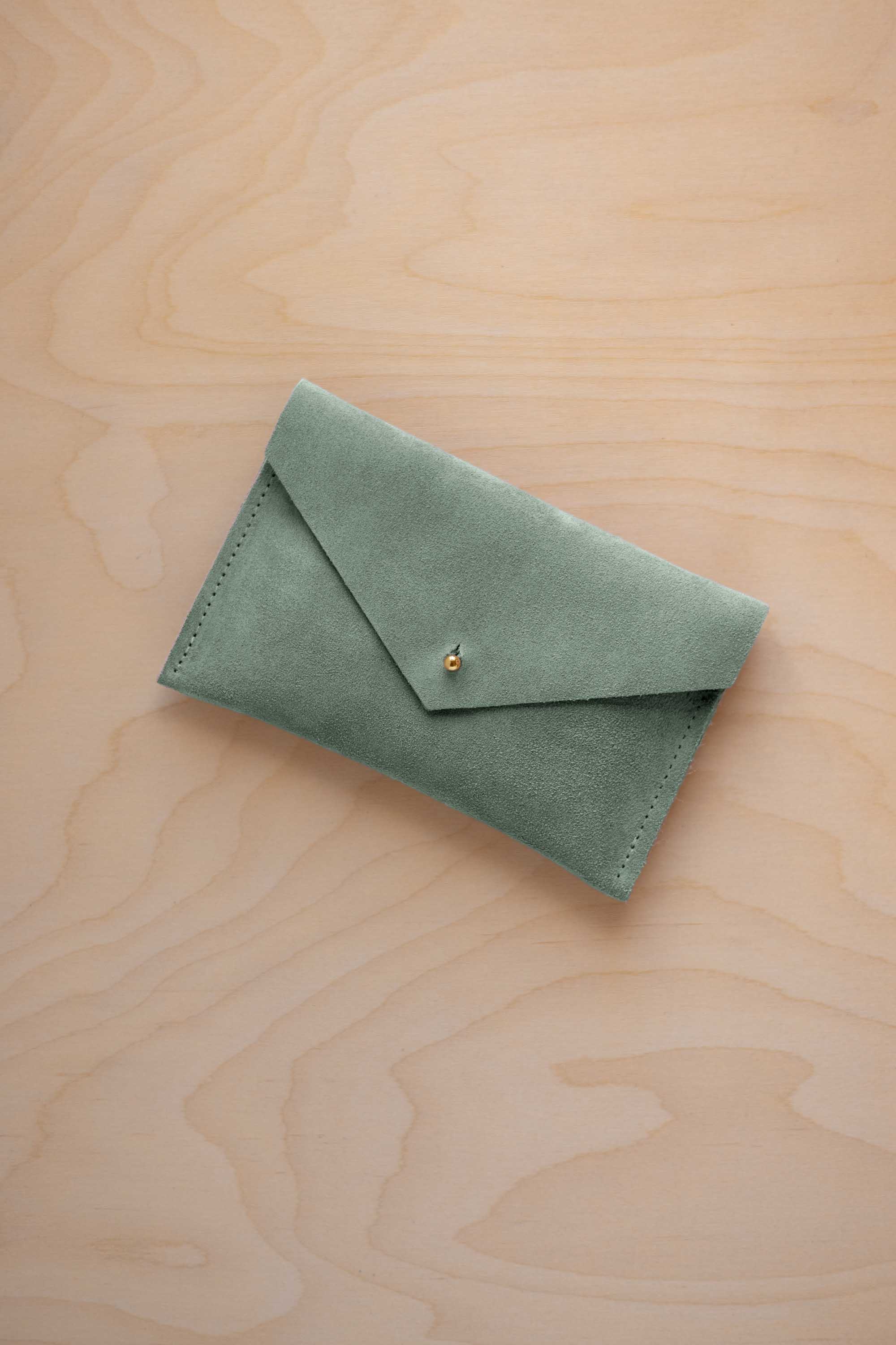 A small eucalyptus green coloured suede envelope-style pouch with a gold button, resting on a light wood grain surface.