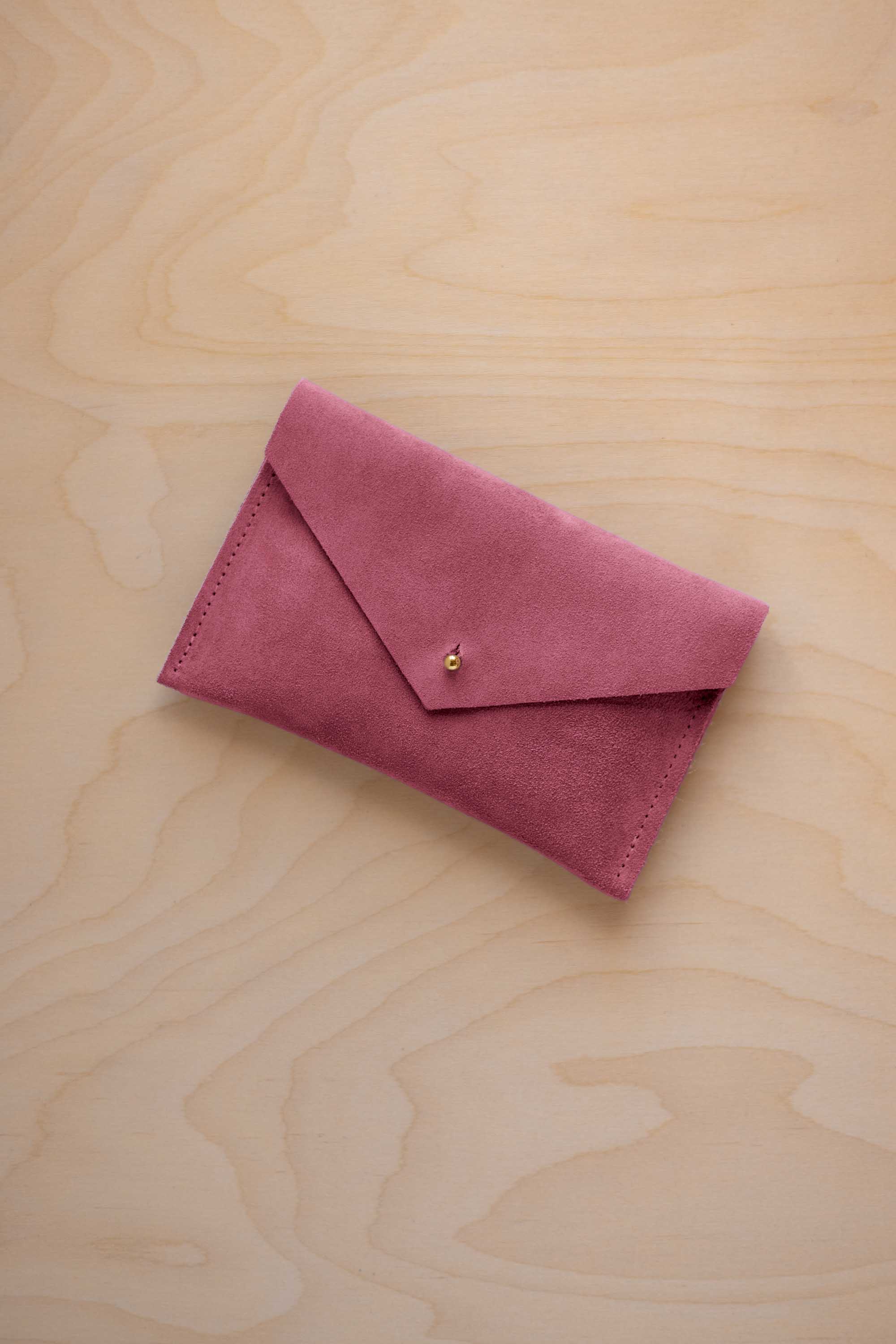 A small faded rose coloured suede envelope-style pouch with a gold button, resting on a light wood grain surface.