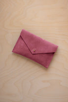 A small faded rose coloured suede envelope-style pouch with a gold button, resting on a light wood grain surface.