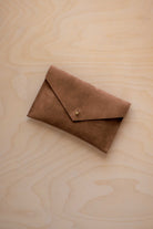 A small mocha brown coloured suede envelope-style pouch with a gold button, resting on a light wood grain surface.