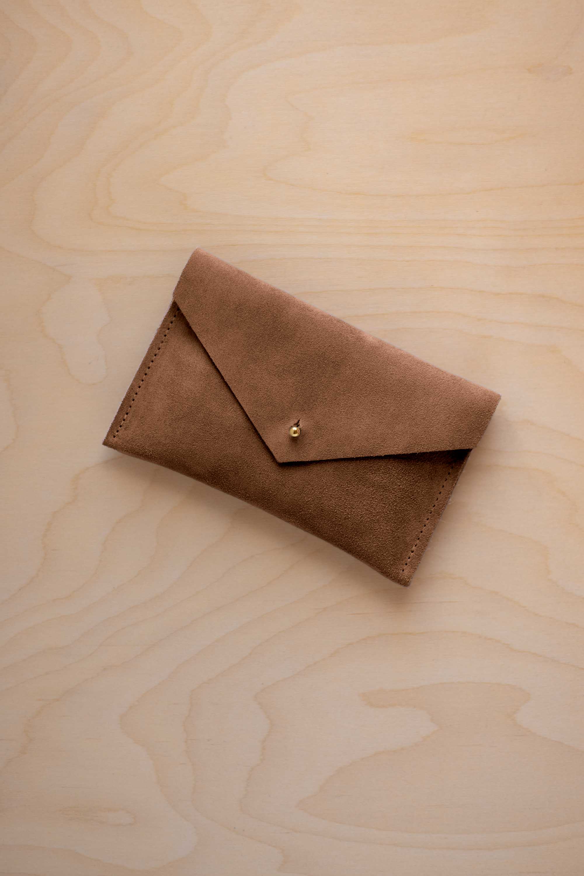 A small mocha brown coloured suede envelope-style pouch with a gold button, resting on a light wood grain surface.