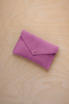 A small peony pink coloured suede envelope-style pouch with a gold button, resting on a light wood grain surface.