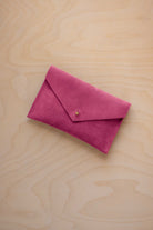 A small raspberry coloured suede envelope-style pouch with a gold button, resting on a light wood grain surface.