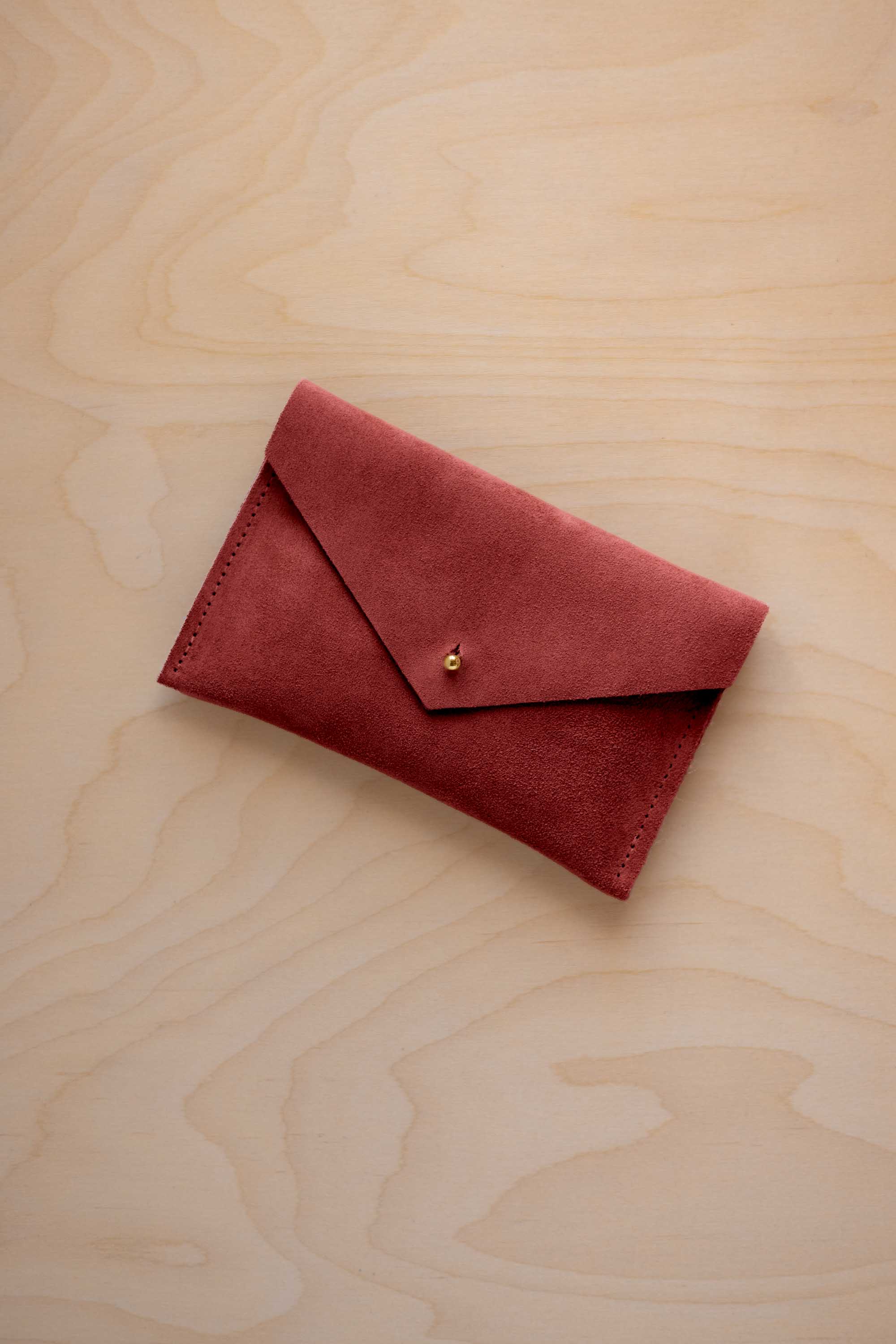 A small rust red coloured suede envelope-style wallet pouch with a gold button, resting on a light wood grain surface.