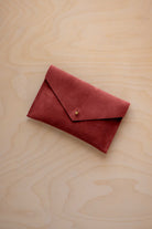 A small rust red coloured suede envelope-style wallet pouch with a gold button, resting on a light wood grain surface.