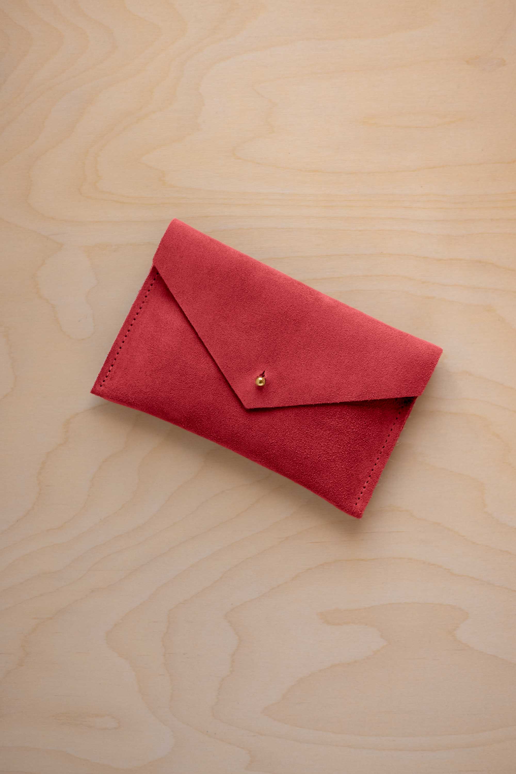 A small scarlet red coloured suede envelope-style pouch with a gold button, resting on a light wood grain surface.