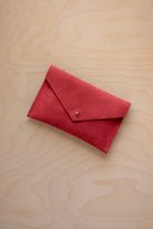 A small scarlet red coloured suede envelope-style pouch with a gold button, resting on a light wood grain surface.