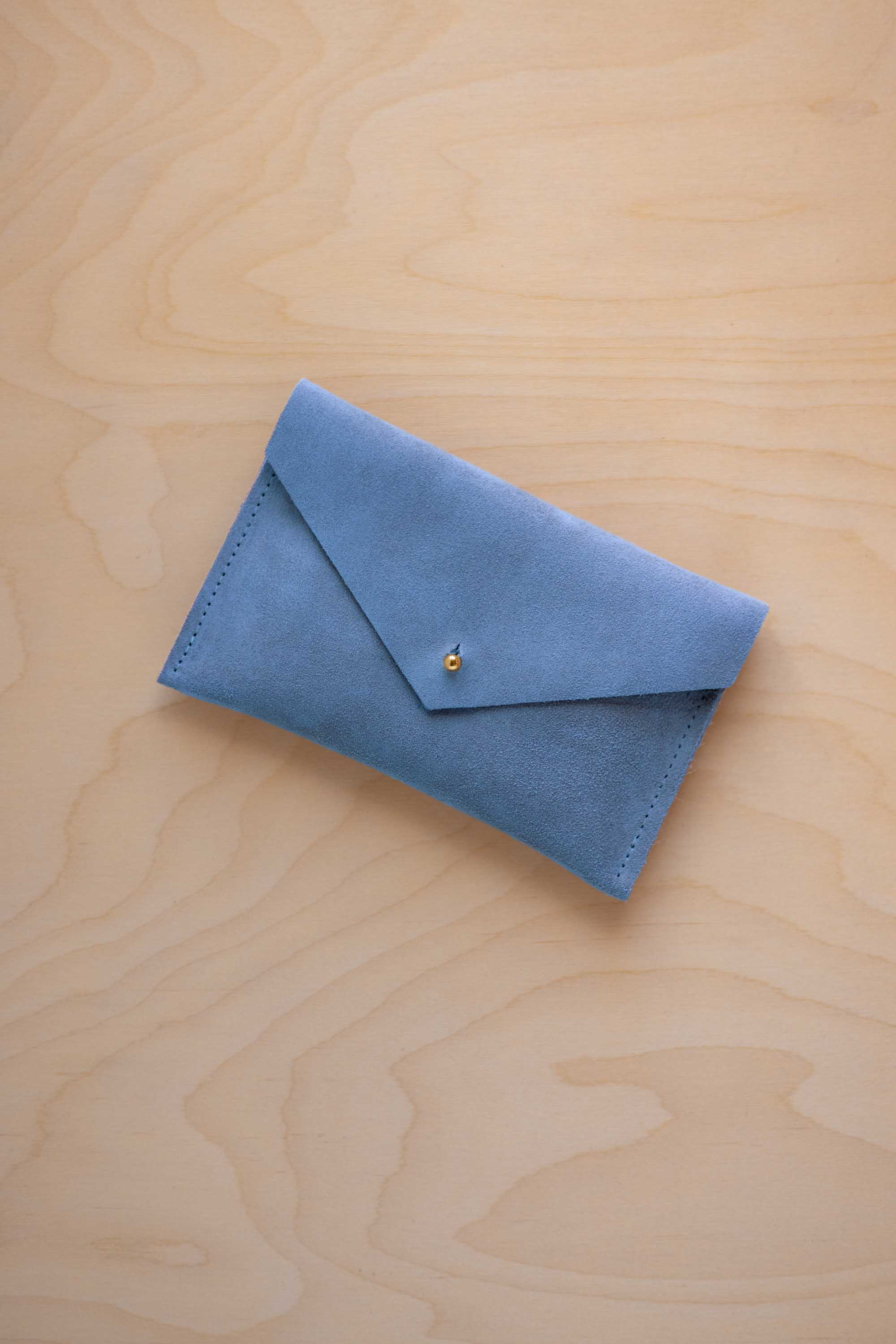 A small sky blue coloured suede envelope-style pouch with a gold button, resting on a light wood grain surface.