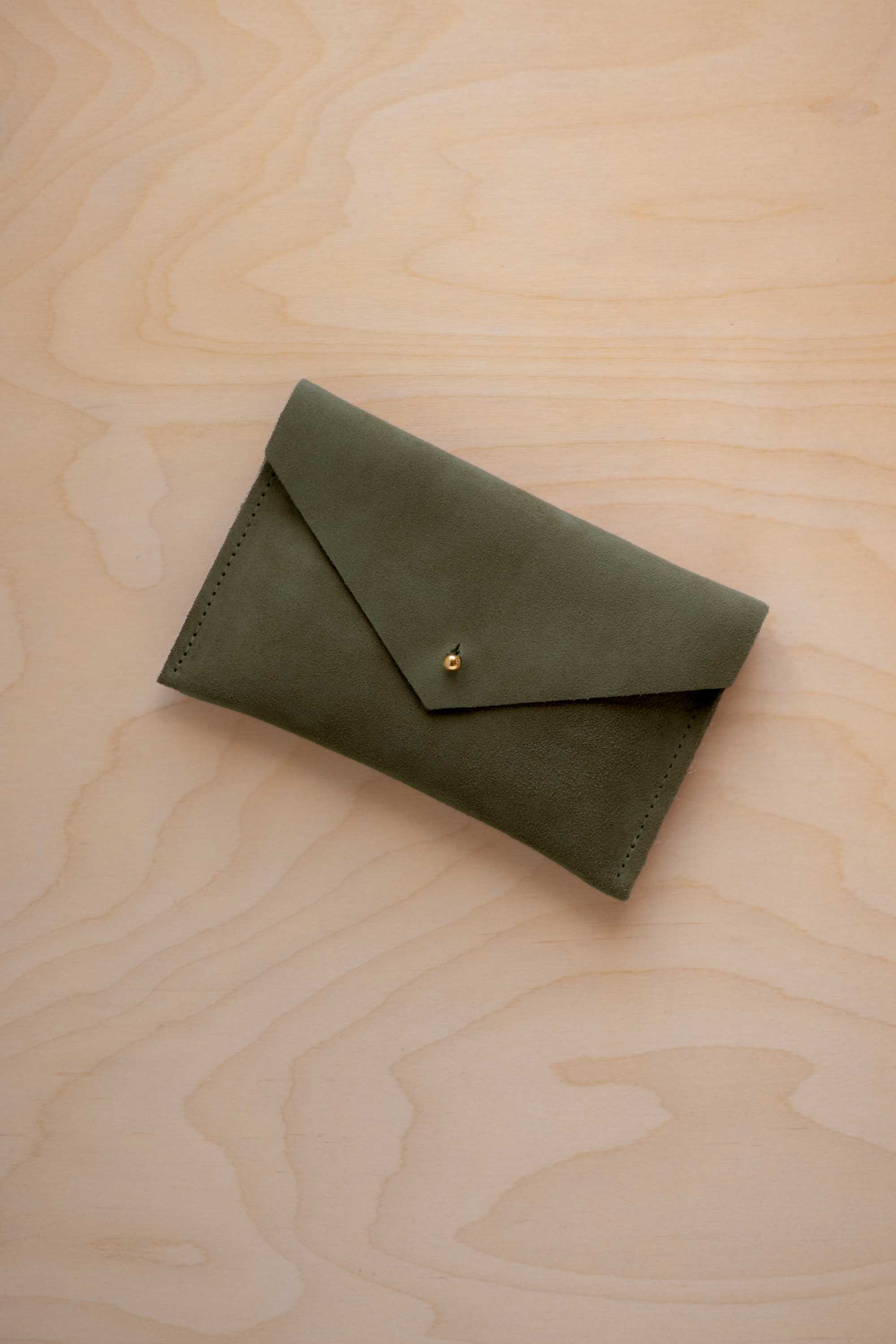 A small soft olive green coloured suede envelope-style pouch with a gold button, resting on a light wood grain surface.
