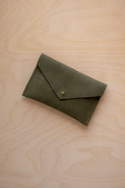 A small soft olive green coloured suede envelope-style pouch with a gold button, resting on a light wood grain surface.
