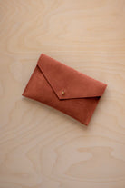A small terracotta coloured suede envelope-style pouch with a gold button, resting on a light wood grain surface.