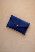 A small ultramarine blue coloured suede envelope-style pouch with a gold button, resting on a light wood grain surface.