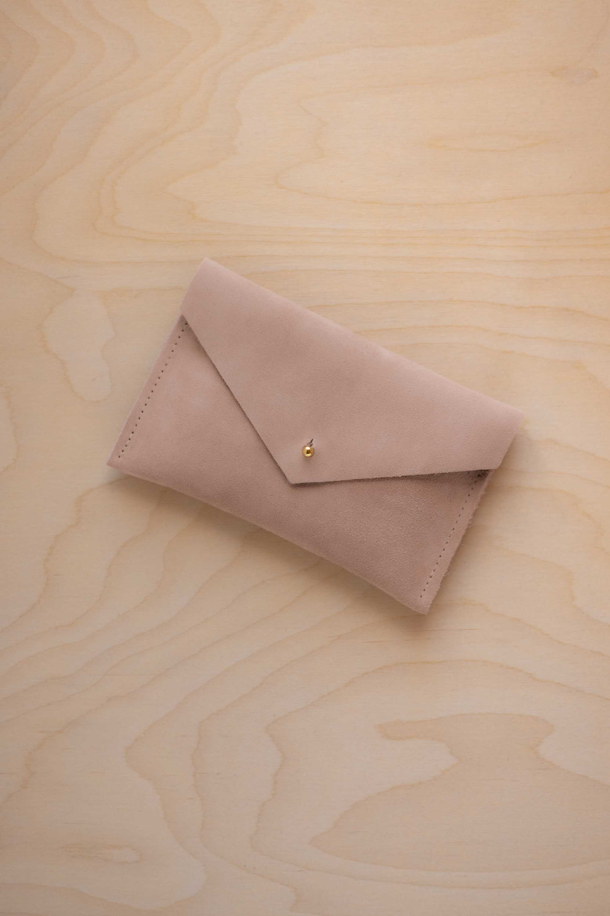 A small warm sand coloured suede envelope-style pouch with a gold button, resting on a light wood grain surface.