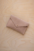 A small warm sand coloured suede envelope-style pouch with a gold button, resting on a light wood grain surface.
