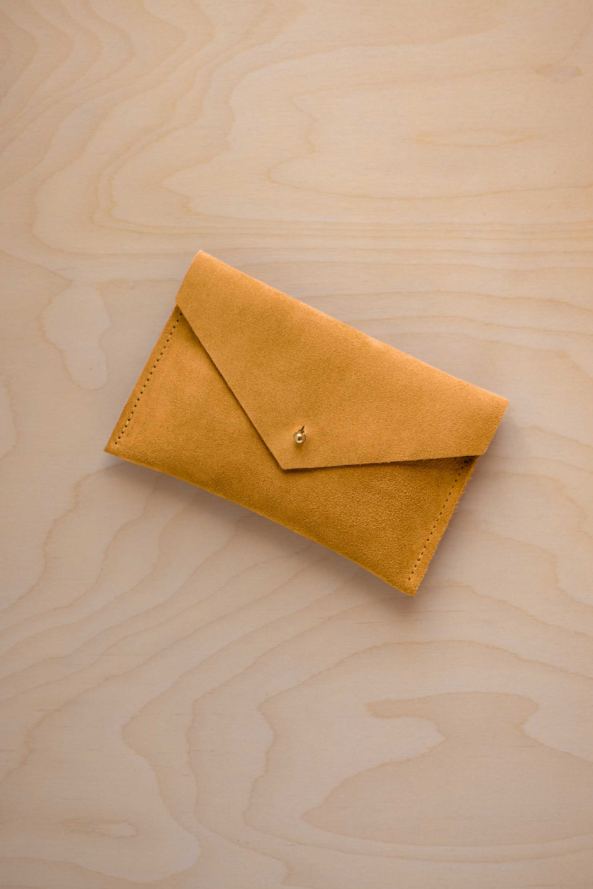 A small golden ochre coloured suede envelope-style pouch with a gold button, resting on a light wood grain surface.