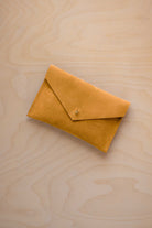A small golden ochre coloured suede envelope-style pouch with a gold button, resting on a light wood grain surface.