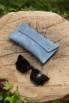 A light blue suede eyeglasses case with gold-tone buttons, shown on a rustic tree stump outdoors with tortoiseshell sunglasses resting in front.