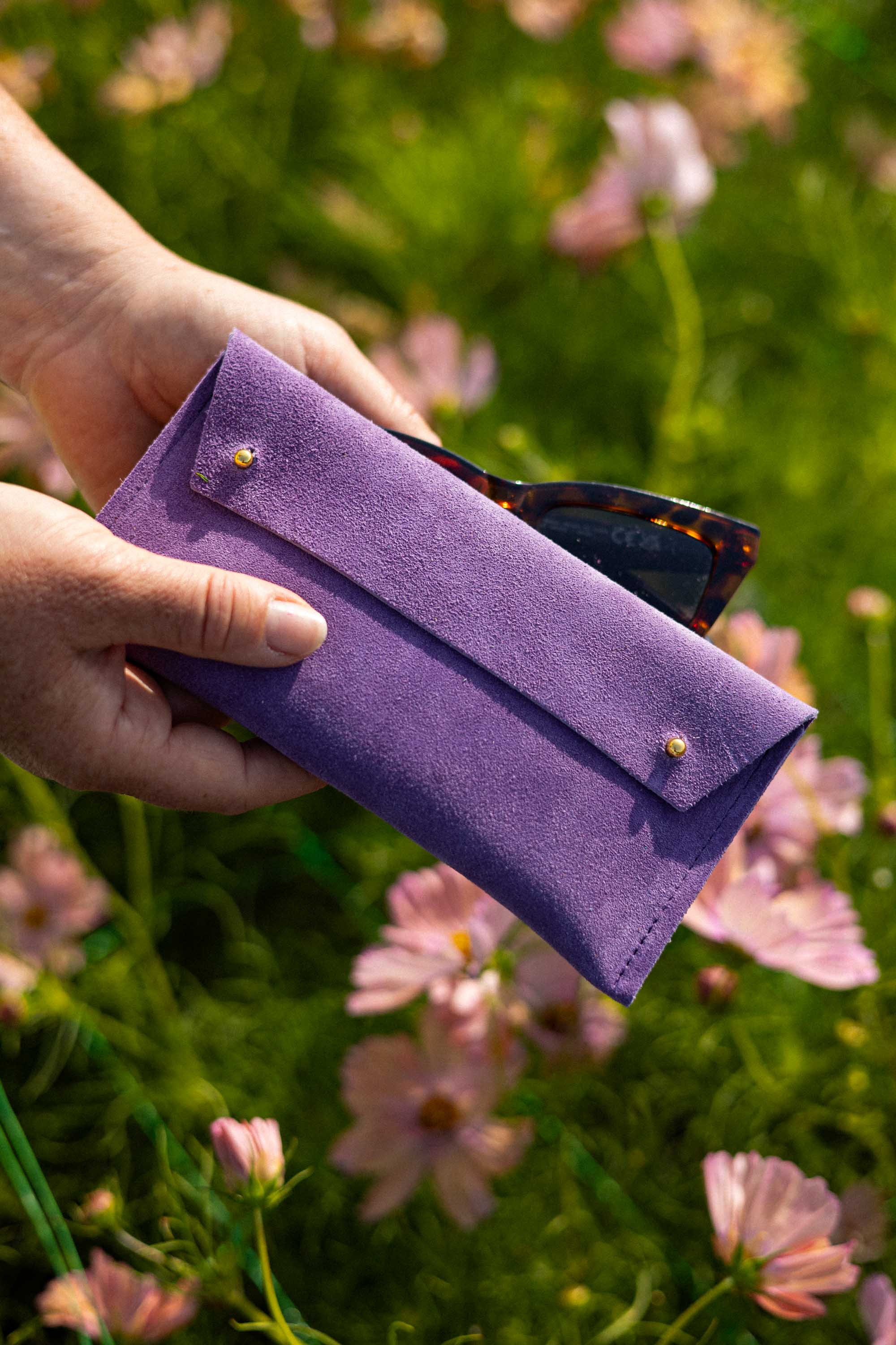 A person's hands holding a purple suede sunglasses case with gold buttons, with tortoiseshell sunglasses partially tucked inside, set against a blurred background of green grass and pink flowers.