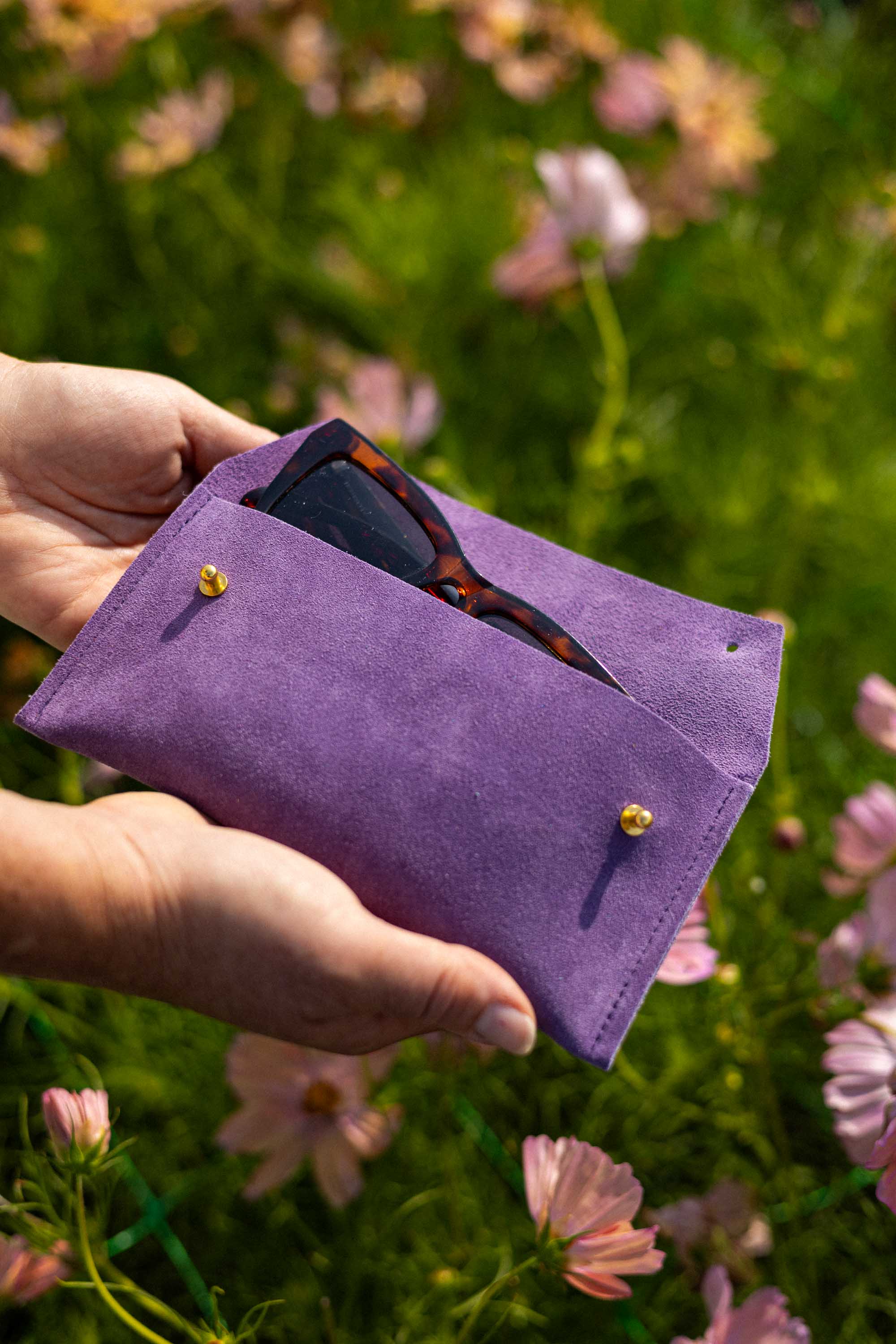 A person's hands holding a purple suede sunglasses pouch with gold buttons, containing tortoiseshell sunglasses, set against a blurred background of green grass and pink flowers.