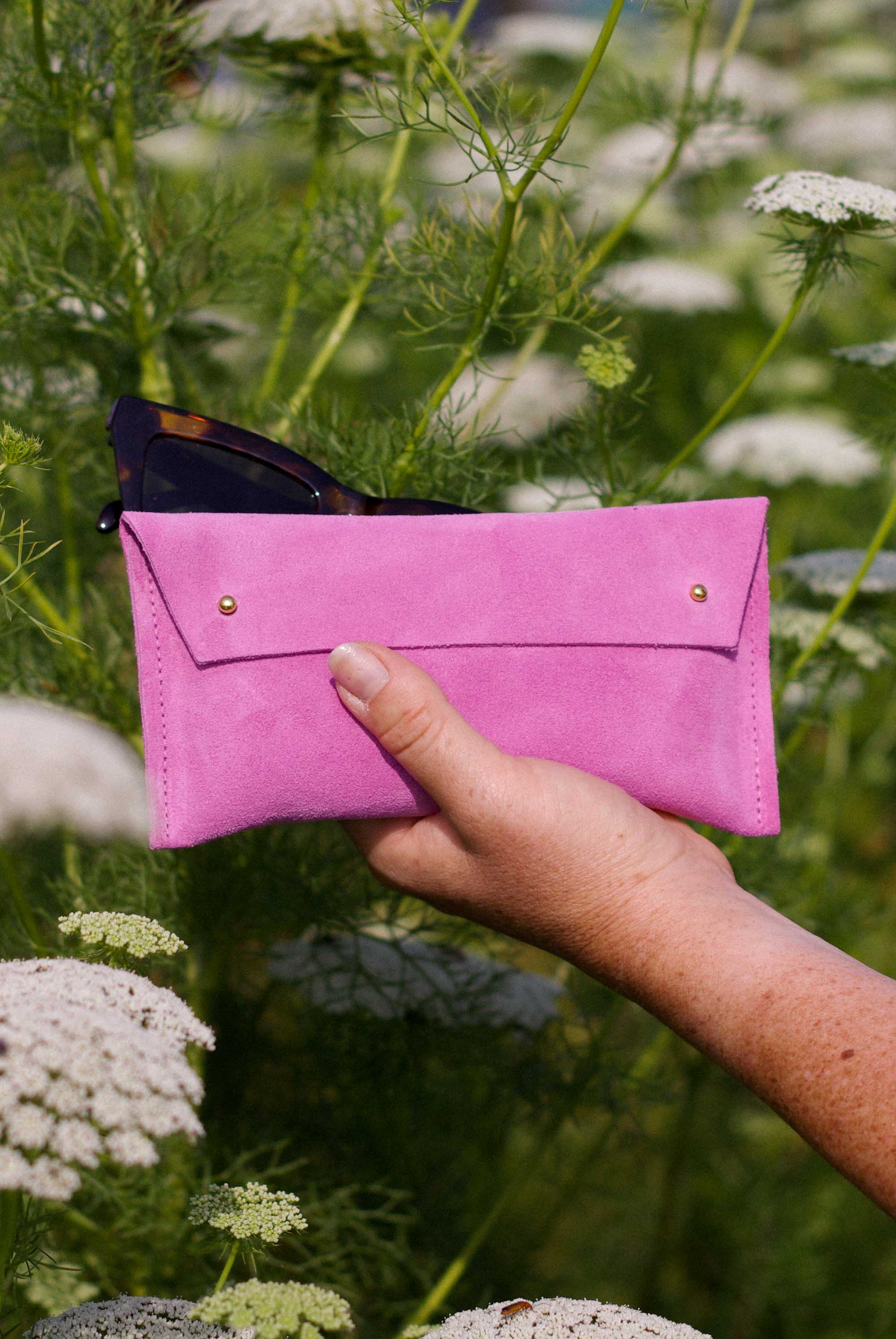 A hand holding a bright pink sunglasses case with gold-tone buttons, set against a blurred background of green plants and white flowers.