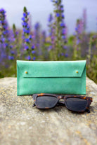 A soft green leather sunglasses case with gold-tone buttons stands on a stone surface outdoors, with tortoiseshell sunglasses placed in front and blurred purple flowers in the background.