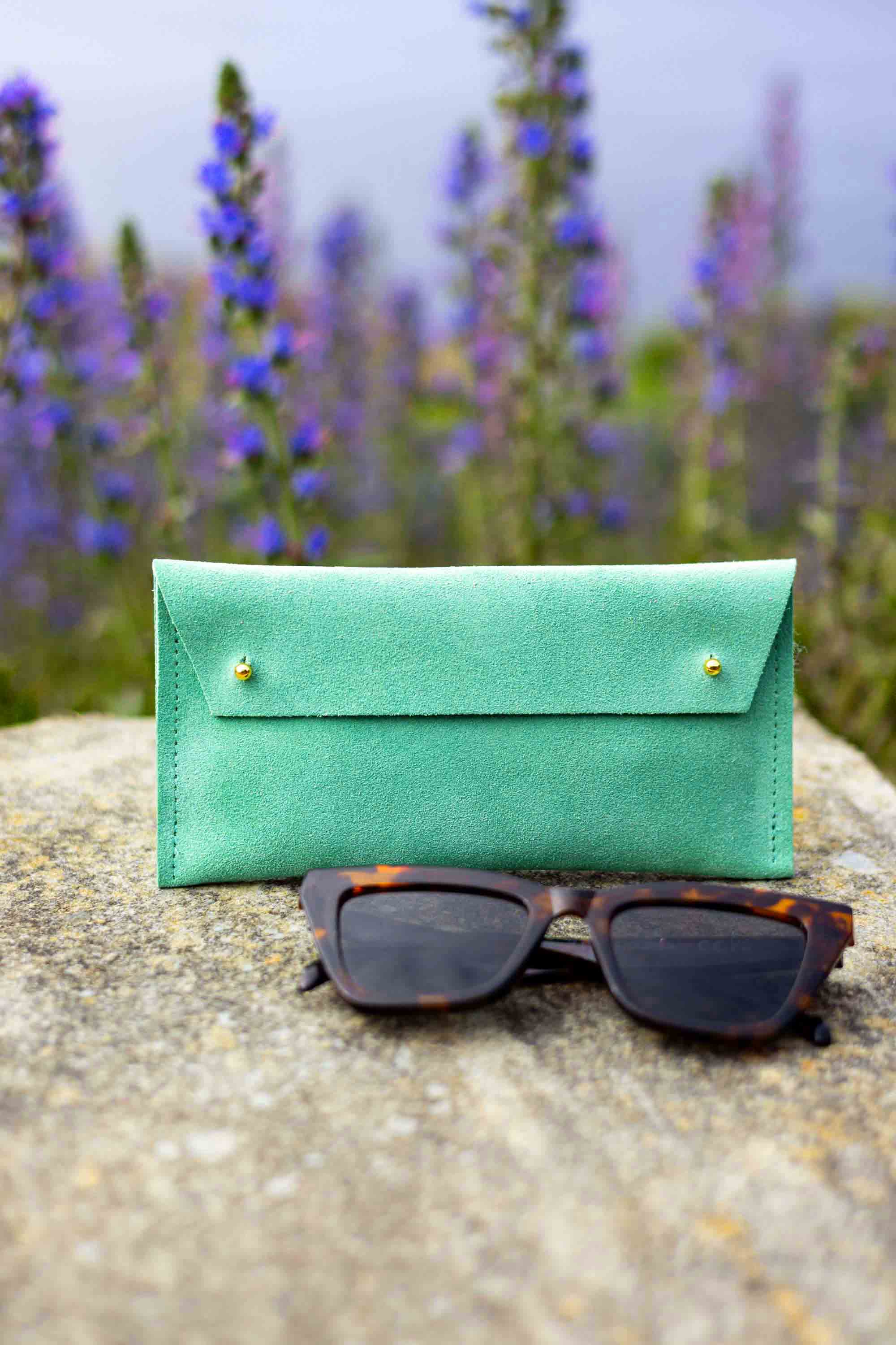 A soft green leather sunglasses case with gold-tone buttons stands on a stone surface outdoors, with tortoiseshell sunglasses placed in front and blurred purple flowers in the background.