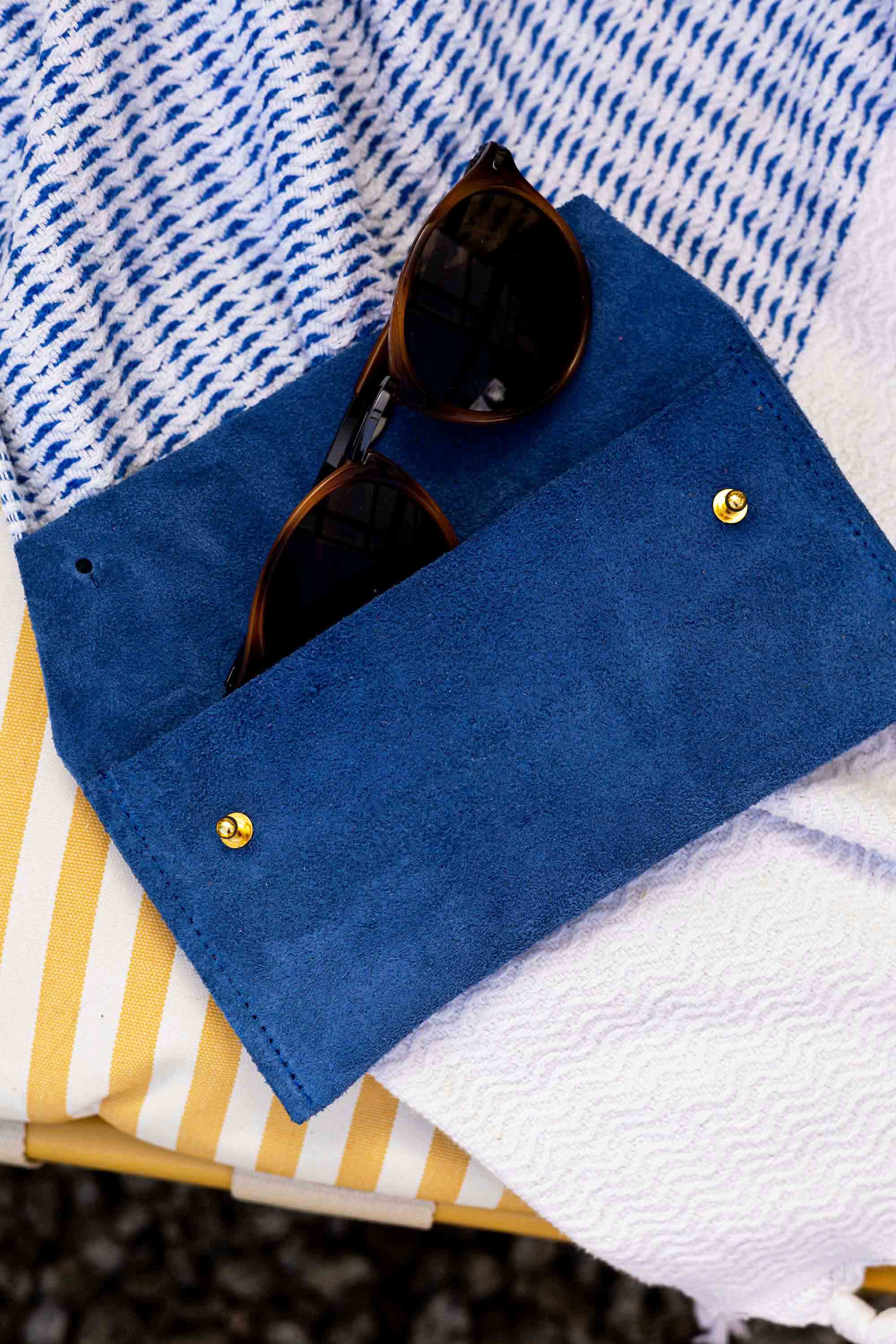 A royal blue blue suede sunglasses case with gold snap buttons, holding a pair of sunglasses, styled on a blue and white striped textile over a yellow and white striped surface.