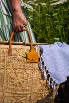 A close-up of a woven straw tote bag with a small mustard yellow suede pouch with gold clip hanging from a tan leather handle, held by a hand outdoors with a white fringed blanket draped over the side.