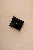 A small black suede pouch with a envelope flap closure and a gold button, placed on a light wood grain surface.