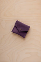 A small dark purple suede pouch with a flap closure and a gold button, placed on a light wood grain surface.