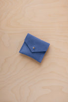 A small cobalt blue suede envelope-style pouch with a gold snap button, placed on a light wood grain surface.