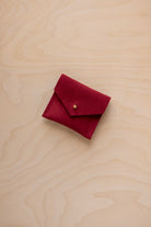A small deep red suede pouch with a flap closure and a gold button, placed on a light wood grain surface.