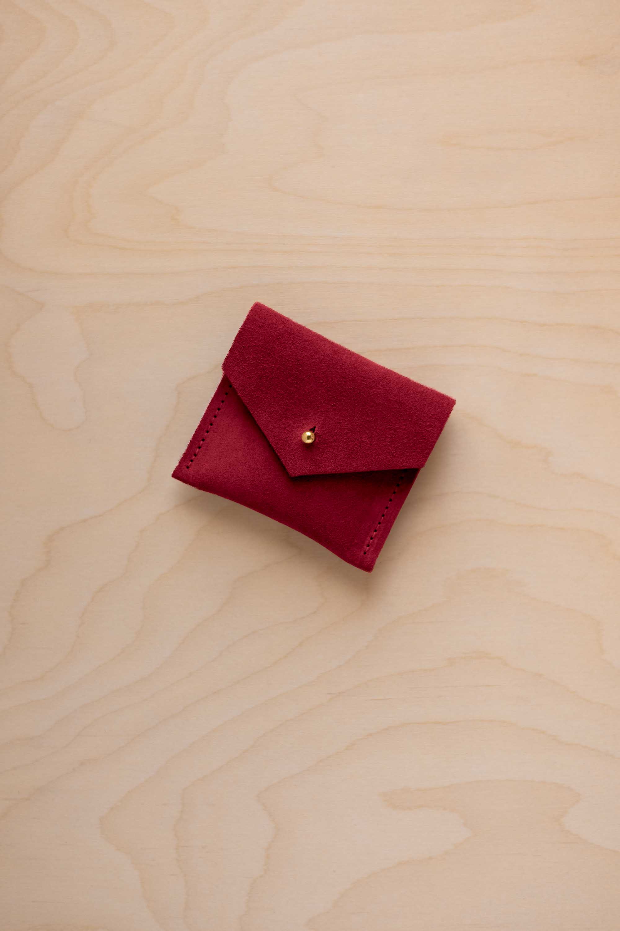 A small deep red suede pouch with a flap closure and a gold button, placed on a light wood grain surface.