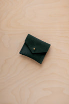 A small dark forest green suede pouch with a envelope flap closure and a gold button, placed on a light wood grain surface.