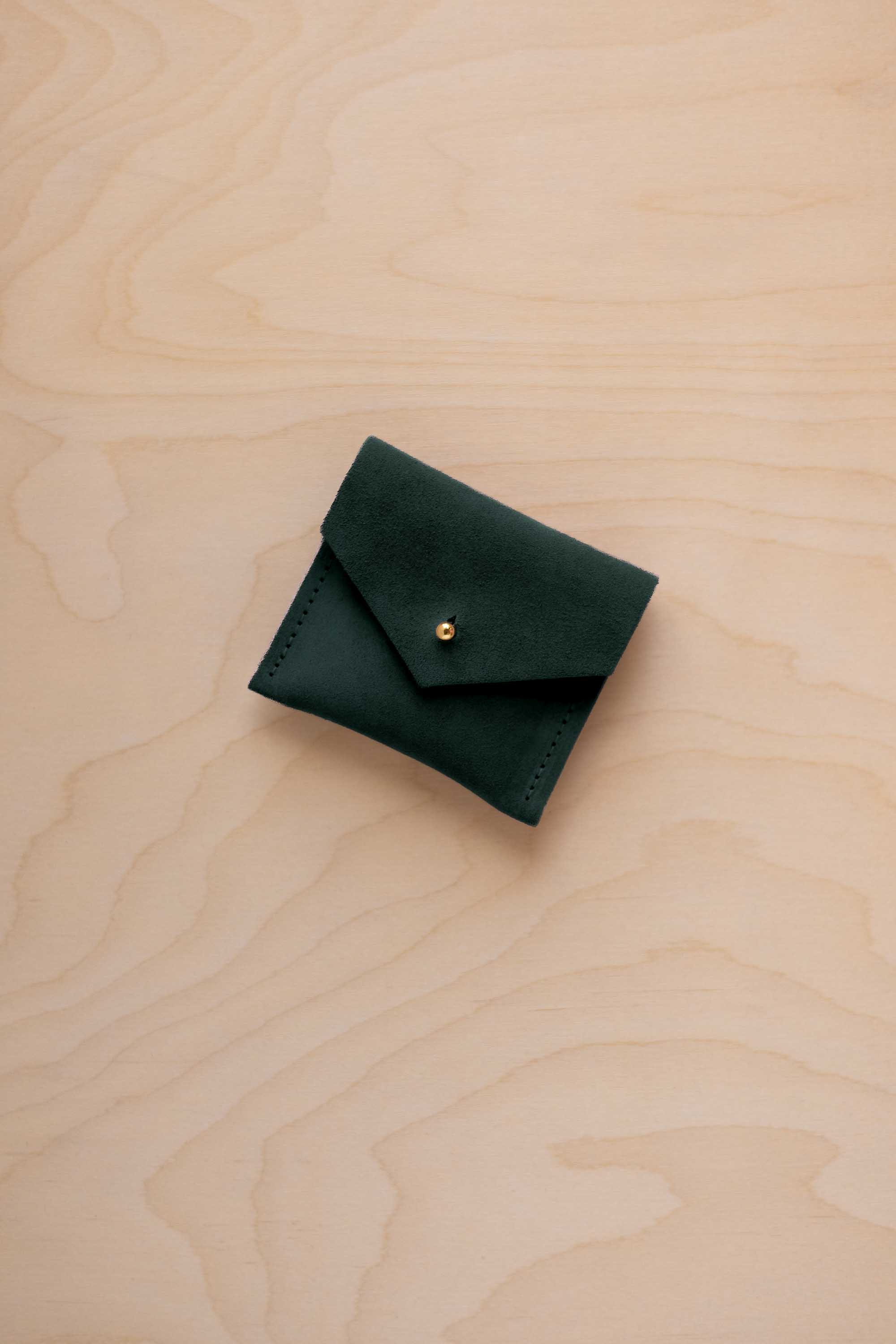 A small dark forest green suede pouch with a envelope flap closure and a gold button, placed on a light wood grain surface.