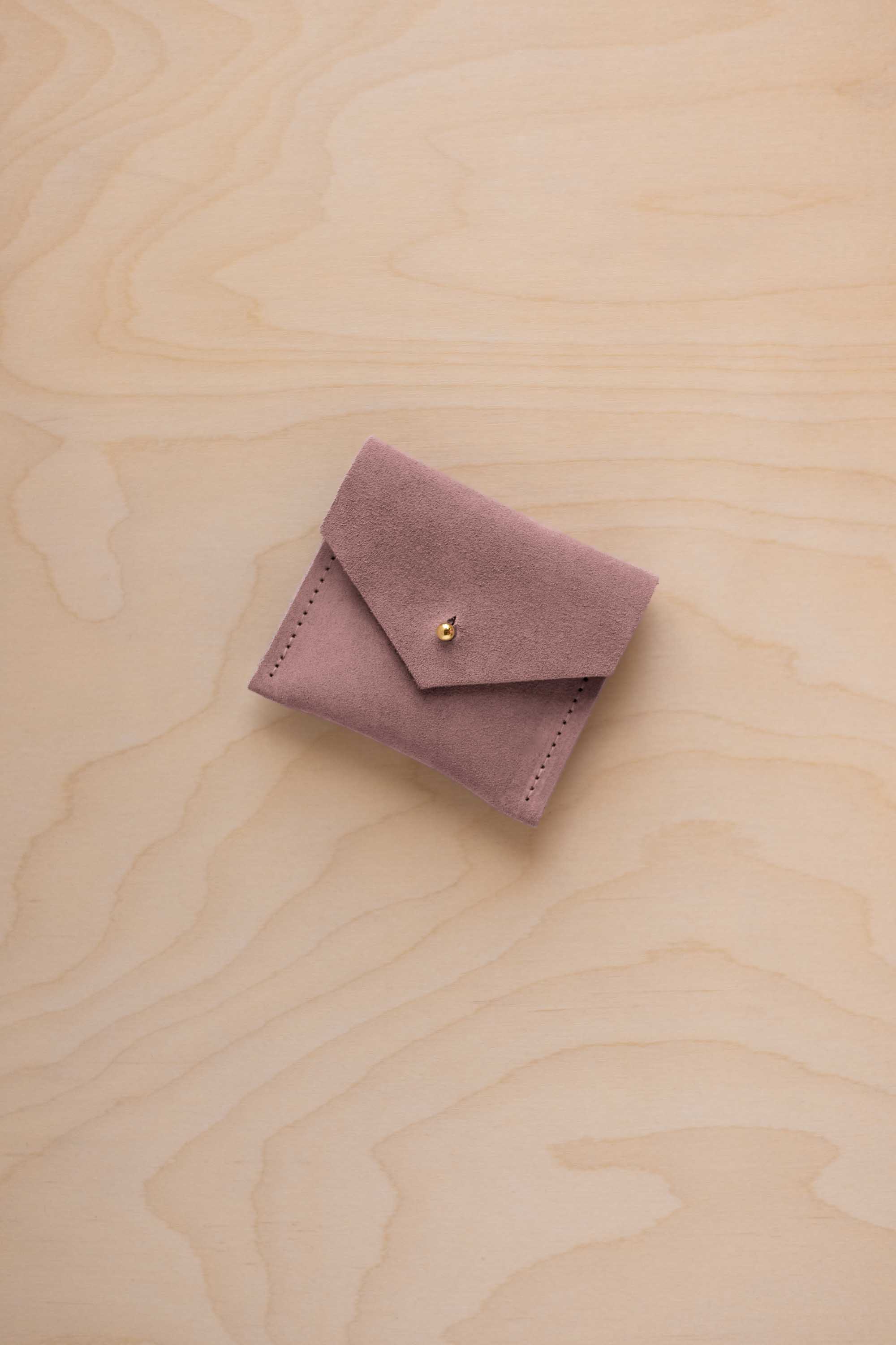 A small dusty mauve suede jewelry pouch with a flap closure and gold button, resting on a light wood grain background.