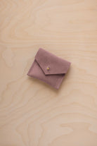 A small dusty mauve suede jewelry pouch with a flap closure and gold button, resting on a light wood grain background.