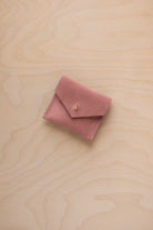 A small dusty pink suede envelope-style pouch with a gold button, resting on a light wood grain surface.