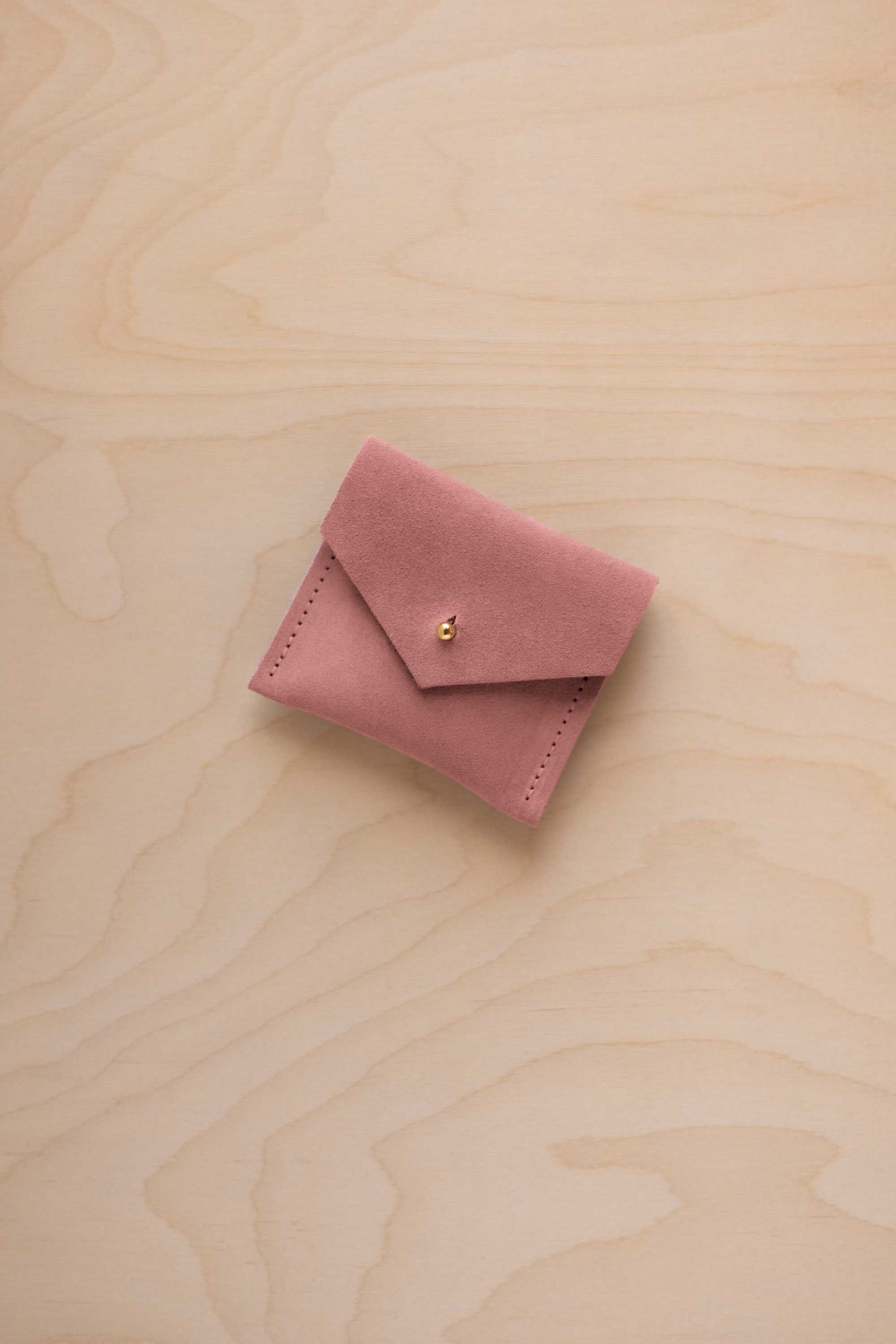 A small dusty pink suede envelope-style pouch with a gold button, resting on a light wood grain surface.