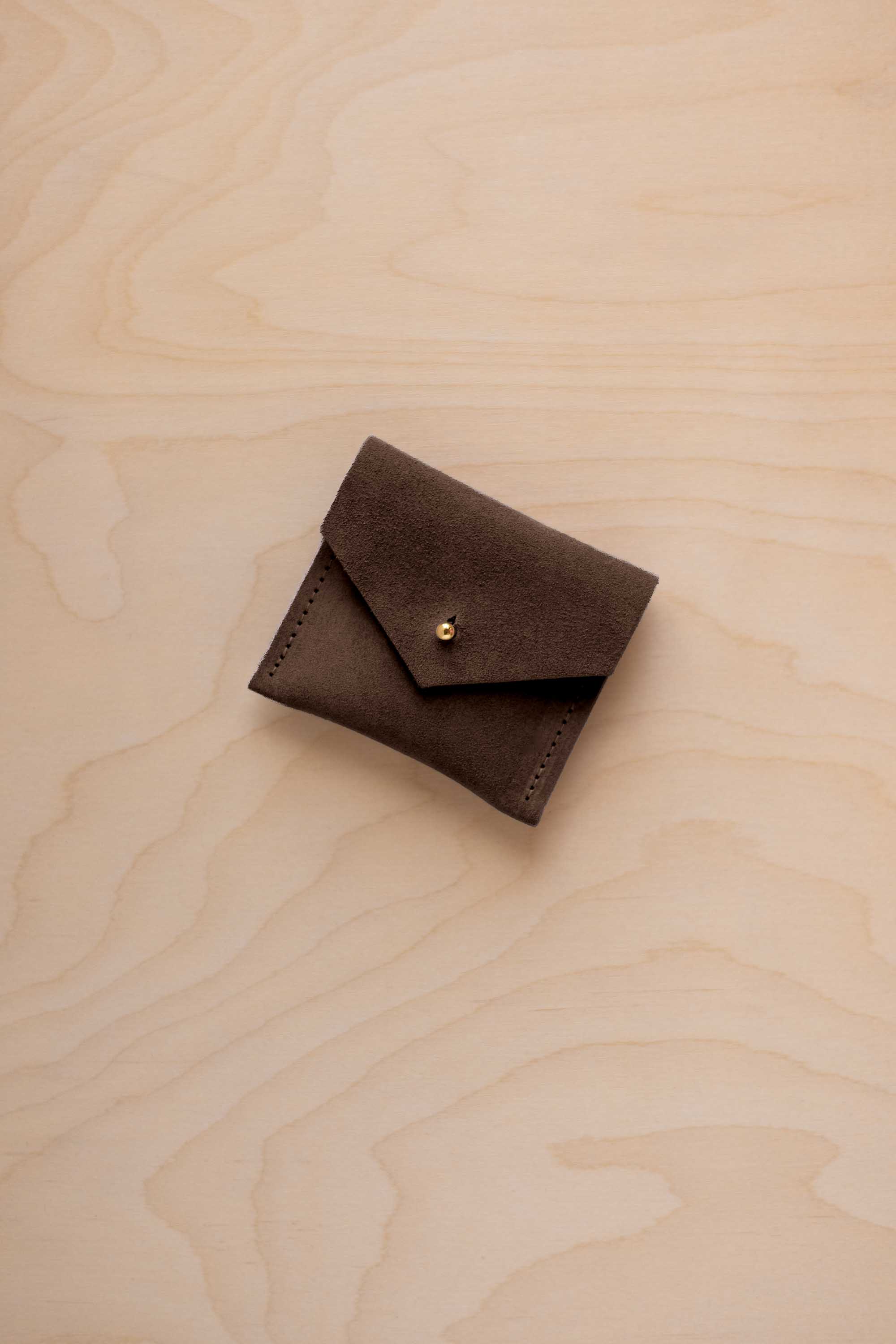 A small dark brown suede jewellery pouch with a flap closure and gold-tone button, placed on a light wood grain surface.